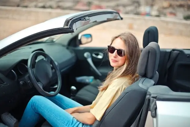 Chica con vaqueros y gafas de sol sentada en el lugar del conductor en un coche de alquiler descapotable